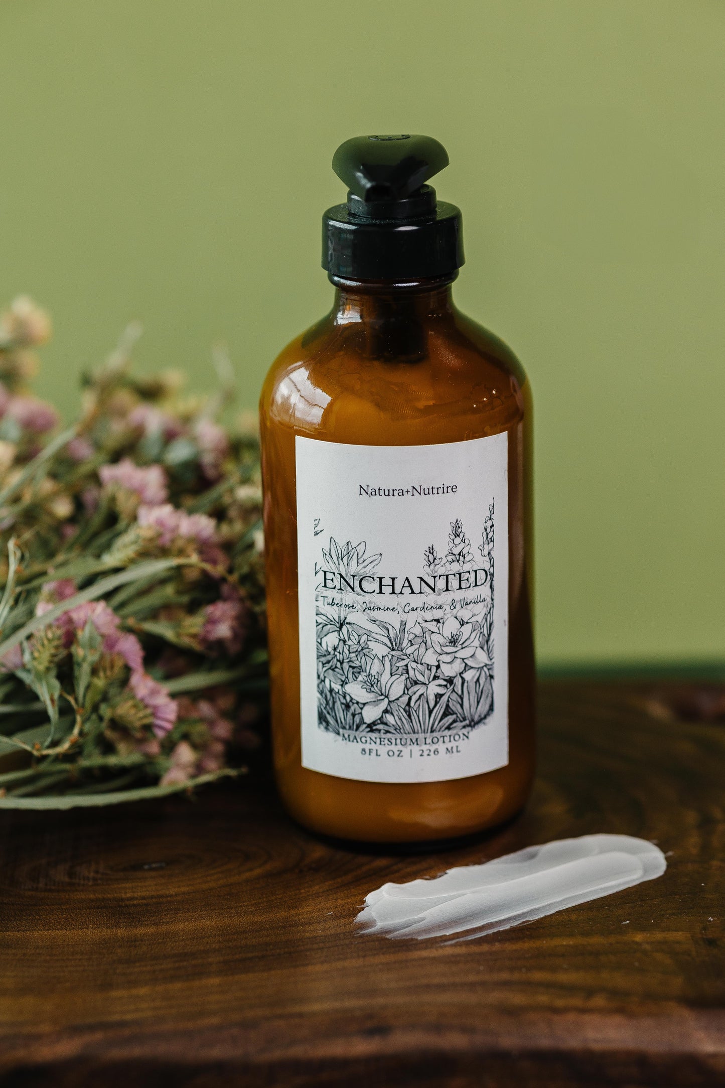 Amber bottle with black pump and 'Enchanted' label on a wooden surface with flowers in the background