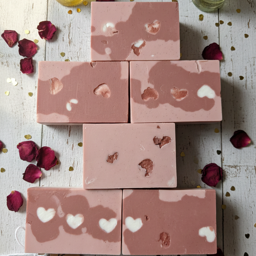 Pink soap bars with heart designs on a wooden surface with rose petals and a 'Valentine's Love' card.