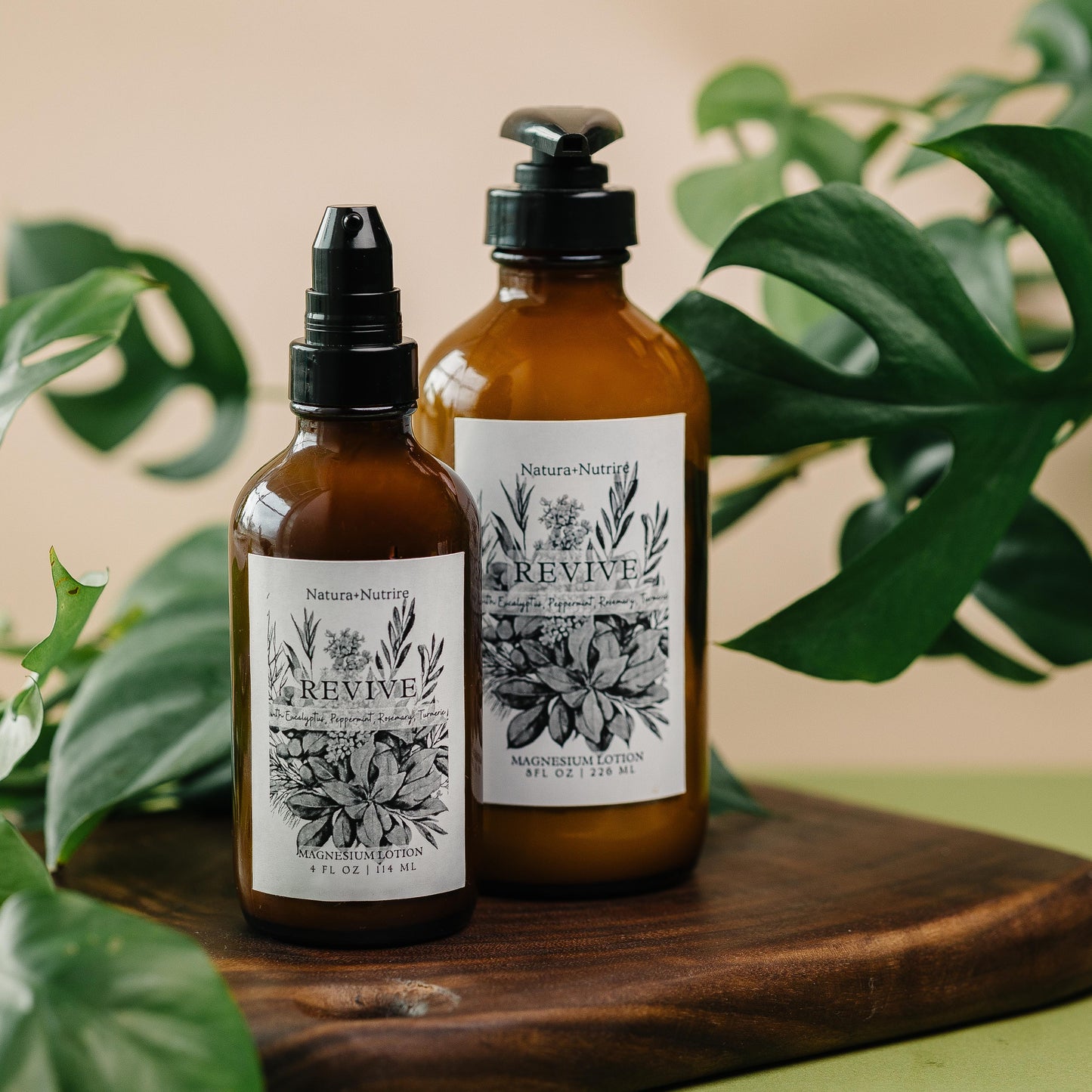 Two brown bottles with black caps on a wooden board surrounded by green leaves.