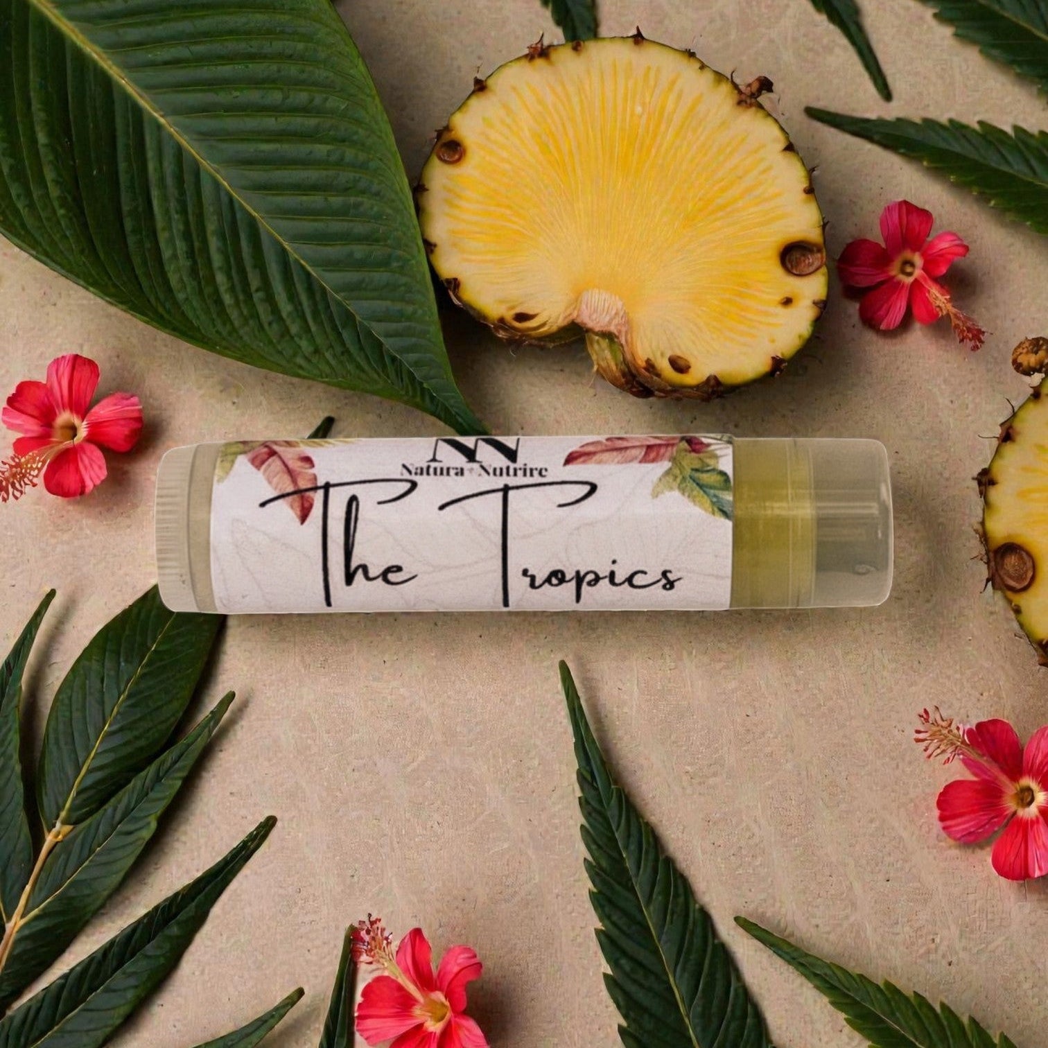 Lip balm labeled 'The Tropics' surrounded by pineapple slices, leaves, and flowers on a beige background.